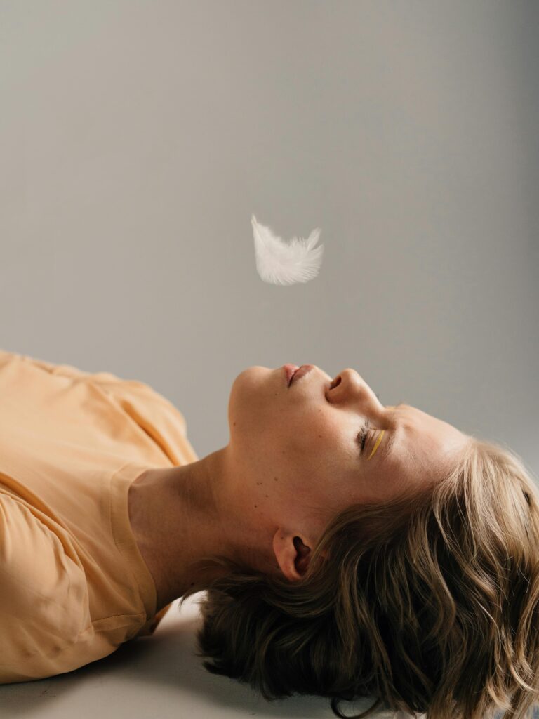 A relaxed woman lies indoors with a feather blowing above her, eyes closed.